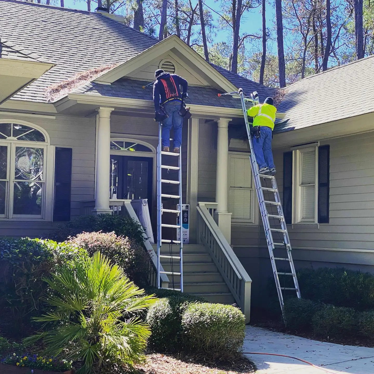 Two A&A crew members on ladders working along the front gutters of a single-story home with palm trees. Gutter cleaning and gutter installation in Bluffton, SC by A&A Island Services